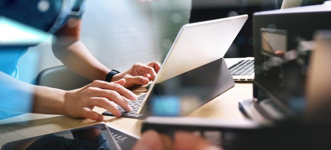 Hands working at computer with person holding cell phone in background